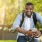 young man smiles with a backpack on and earphones in as he sits on his bicycle in the park